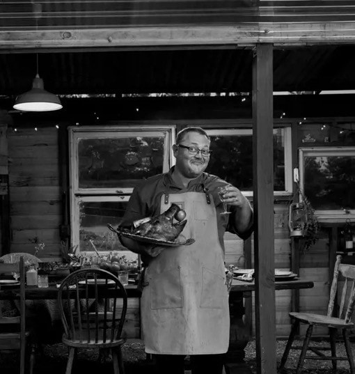 Portrait of the owner of Fork It Farm in Tasmania, standing proudly outside a rustic dining area. He wears a leather apron and smiles warmly, holding a smoked cut of meat on a carving board. The background features a charming wooden cabin with visible dining tables, adding to the farmstay experience in Tasmania.